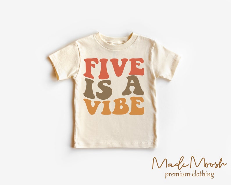 May include: A cream colored t-shirt with the text "FIVE IS A VIBE" in retro-style font with orange, red, and brown colors. The text is centered on the shirt.