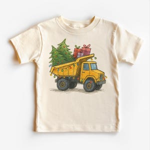 May include: Cream-colored t-shirt featuring a yellow dump truck carrying a green Christmas tree and wrapped gifts. The truck is decorated with colorful lights, suggesting a festive holiday theme. The design is a hand-drawn style.