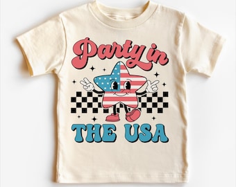 May include: A cream-coloured t-shirt with a retro-style graphic of a cartoon star wearing an American flag design. The text "Party in the USA" is printed below the star.