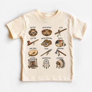 May include: A cream-colored short-sleeve t-shirt featuring a grid of twelve illustrations of cultural items labeled Pottery, Woven Basket, Flute, Clay Pipe, Beaded Moccasins, Shield, Hand Drum, Spear, Tomahawk, Weaving, Dreamcatcher, and Headdress.
