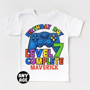 May include: White t-shirt with a colorful graphic of a video game controller and the text "Birthday Boy Level 7 Complete Maverick".