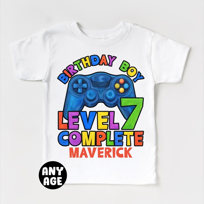 May include: White t-shirt with a colorful graphic of a video game controller and the text "Birthday Boy Level 7 Complete Maverick".