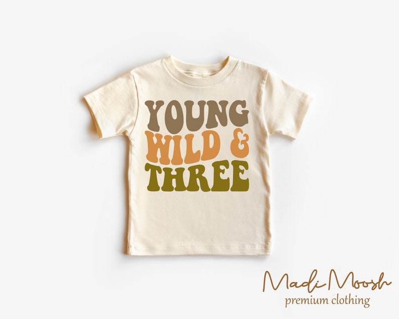 May include: A cream colored t-shirt with the text "YOUNG WILD & THREE" in brown, orange, and green lettering. The shirt is a toddler size and has short sleeves.