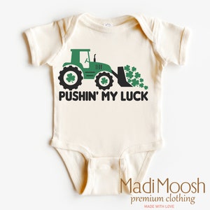 Może przedstawiać: Kremowe body dla niemowląt z zieloną grafiką traktora i napisem "PUSHIN' MY LUCK". Traktor ma czarne koła z detalami koniczyny i wysypuje stos koniczyny. Nazwa marki "Madi Moosh" jest wydrukowana na dole.