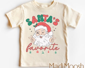 Puede incluir: Camiseta infantil color crema con un diseño retro de Papá Noel. El diseño presenta el texto "SANTA'S favorite" en rojo y verde, con Papá Noel guiñando un ojo y haciendo el signo de la paz. Iconos navideños debajo.