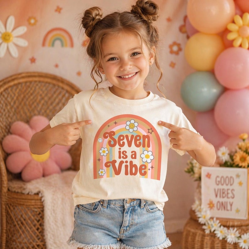 May include: A young girl wearing a cream-colored t-shirt with a retro rainbow design that says "Seven is a Vibe." The shirt is paired with denim shorts. The background includes balloons, flowers, and a sign that says "Good Vibes Only."