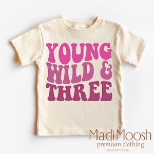 May include: A cream colored t-shirt with the text "Young Wild & Three" in pink and purple ombre lettering. The text is in a retro font style. The shirt is made by Madi Moosh premium clothing.