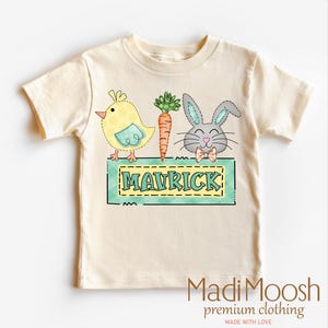 May include: A cream-colored t-shirt featuring a cartoon design with a yellow chick, an orange carrot, and a gray bunny with a bow tie. The name "MAVRICK" is displayed on a green and yellow sign. The shirt is from MadiMoosh premium clothing.
