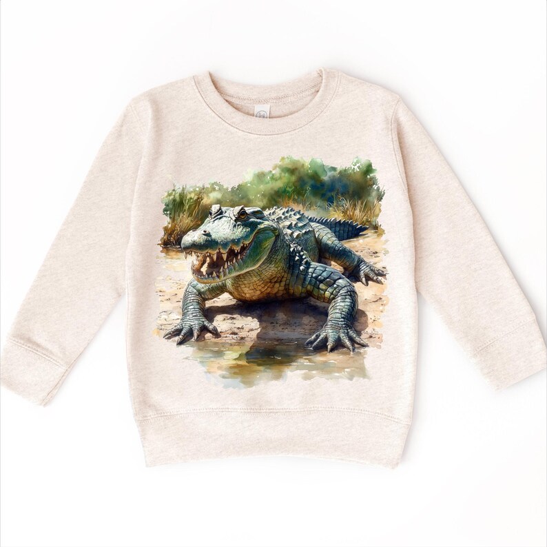 K&ouml;nnte beinhalten: Ein beiger Pullover mit einer Aquarell-Illustration eines Alligators auf der Vorderseite. Der Alligator ist gr&uuml;n und braun mit einem wei&szlig;en Bauch und wird in einer nat&uuml;rlichen Umgebung gezeigt.