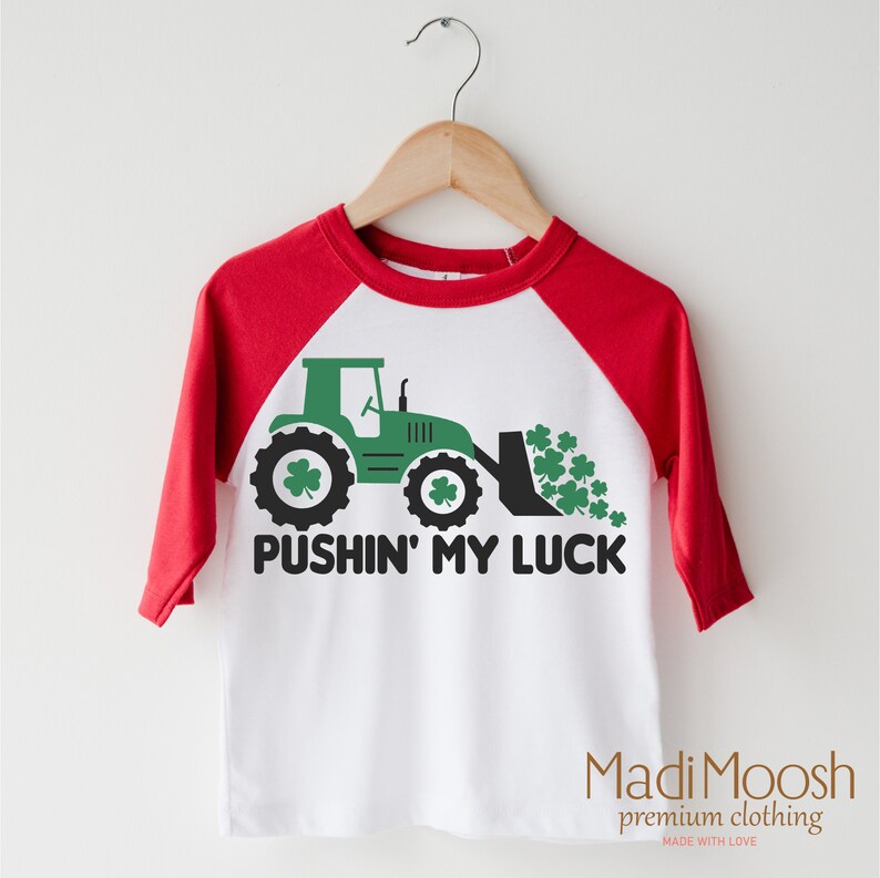 Może przedstawiać: Biało-czerwona koszulka raglan z zieloną grafiką traktora i napisem "PUSHIN' MY LUCK". Traktor ma koniczyny na kołach i w łyżce. Koszulka wisi na drewnianym wieszaku.