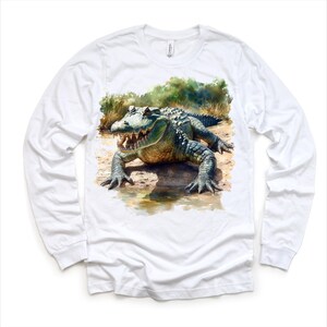 K&ouml;nnte beinhalten: Wei&szlig;es Langarm-T-Shirt mit einer Aquarell-Illustration eines Alligators in einer Sumpflandschaft.