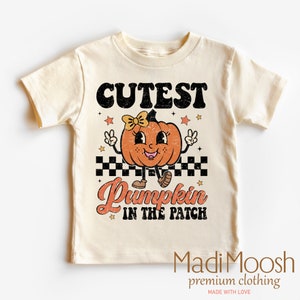 May include: Cream-colored t-shirt featuring a retro-style pumpkin graphic with a bow, peace signs, and the text "Cutest Pumpkin in the Patch." The design includes stars and a checkered pattern, perfect for fall.