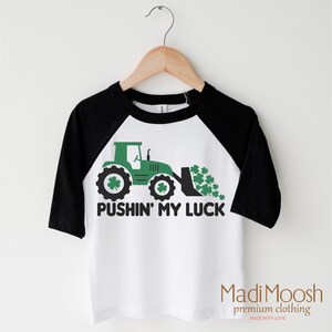 Może przedstawiać: Biała i czarna koszulka z rękawami raglan z zieloną grafiką traktora i napisem "PUSHIN' MY LUCK". Traktor ma detale koniczyny, a nazwa marki "Madi Moosh" znajduje się na dole.