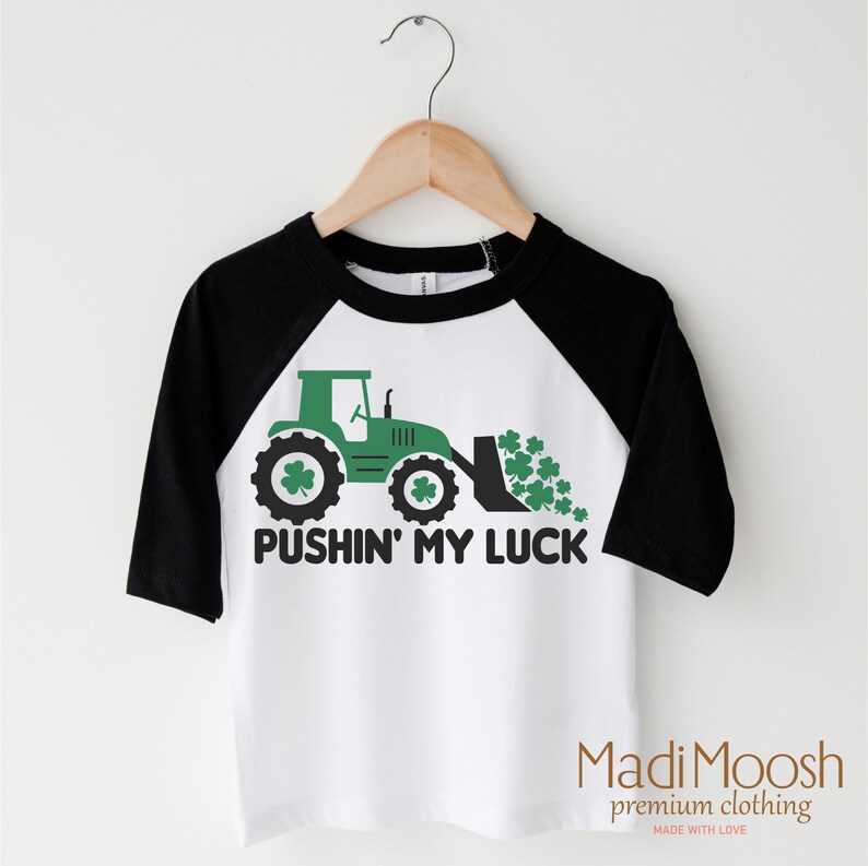 Może przedstawiać: Biała i czarna koszulka z rękawami raglan z zieloną grafiką traktora i napisem "PUSHIN' MY LUCK". Traktor ma detale koniczyny, a nazwa marki "Madi Moosh" znajduje się na dole.