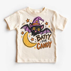 May include: A cream-colored short-sleeve t-shirt featuring a crayon-style illustration of a black bat wearing a purple witch hat. The bat is flying near a yellow crescent moon and orange stars, with the text "BATTY FOR CANDY" printed in orange.