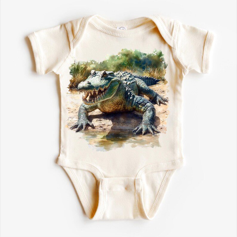 K&ouml;nnte beinhalten: Ein wei&szlig;er Baby-Strampler mit einer Aquarell-Illustration eines Alligators in einer sumpfigen Umgebung.