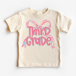 May include: A cream-colored t-shirt featuring the words "Third Grade" in a pink, bubble-style font, accented with a colorful bow design. The bow has pink, blue, and yellow details. The shirt is made of soft material and is suitable for children.