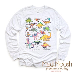 May include: White long-sleeve t-shirt with a colorful dinosaur graphic print. The print features various dinosaurs with their names written underneath. The shirt has a crew neck and long sleeves. The text "Madi Moosh premium clothing MADE WITH LOVE" is printed at the bottom of the shirt.