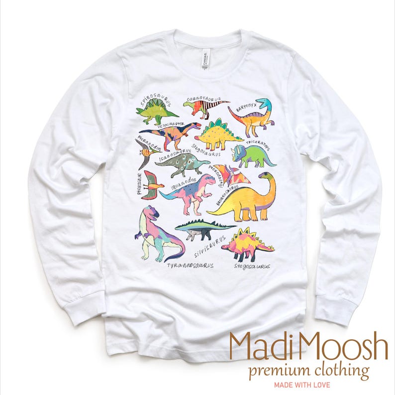 May include: White long-sleeve t-shirt with a colorful dinosaur graphic print. The print features various dinosaurs with their names written underneath. The shirt has a crew neck and long sleeves. The text "Madi Moosh premium clothing MADE WITH LOVE" is printed at the bottom of the shirt.
