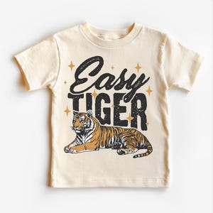 May include: A beige short-sleeved t-shirt with the text "Easy Tiger" in black lettering and a stylized illustration of a tiger in brown and orange. The text is surrounded by small gold stars.
