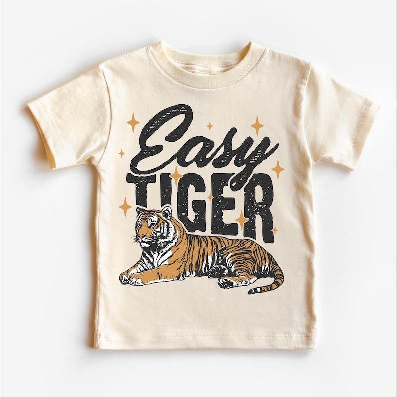 Op de afbeelding: Een beige T-shirt met korte mouwen met de tekst "Easy Tiger" in zwarte letters en een gestileerde illustratie van een tijger in bruin en oranje. De tekst is omringd door kleine gouden sterren.
