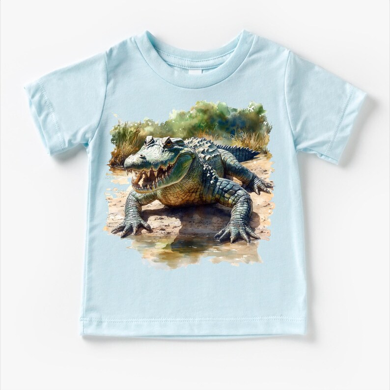 K&ouml;nnte beinhalten: Ein hellblaues Baby-T-Shirt mit einer Aquarell-Illustration eines Alligators auf einem gr&uuml;n-braunen Hintergrund.