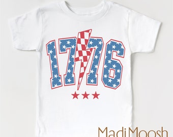 May include: White baby t-shirt featuring a red, white and blue 1776 graphic. The graphic includes a lightning bolt with stars and a chequered pattern. The text "Madi Moosh premium clothing" is printed below the graphic.