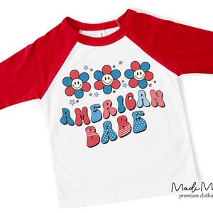 May include: White and red raglan style t-shirt with three-quarter sleeves. The shirt features a graphic with three smiling flower faces and the words "AMERICAN BABE" in red, white, and blue. The shirt is from the brand "Madi Moosh premium clothing."