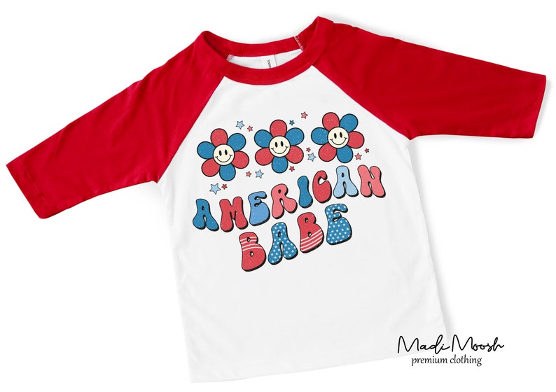 May include: White and red raglan style t-shirt with three-quarter sleeves. The shirt features a graphic with three smiling flower faces and the words "AMERICAN BABE" in red, white, and blue. The shirt is from the brand "Madi Moosh premium clothing."
