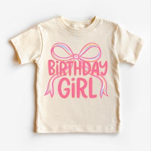 May include: A cream-colored t-shirt featuring the text "BIRTHDAY GIRL" in pink, with a pink bow and pastel-colored ribbons. The shirt is short-sleeved and suitable for children.
