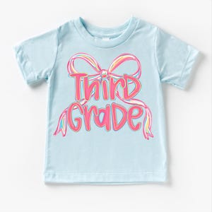 May include: A light blue t-shirt with the words "Third Grade" in pink, outlined in a darker pink. The text is designed with a bow graphic in the background, with rainbow-colored accents.