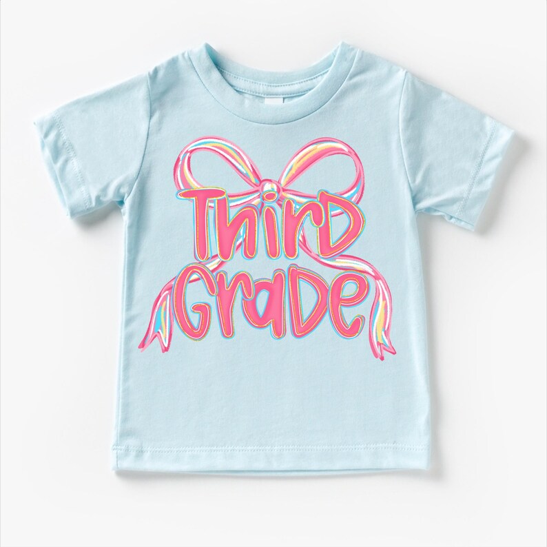 May include: A light blue t-shirt with the words "Third Grade" in pink, outlined in a darker pink. The text is designed with a bow graphic in the background, with rainbow-colored accents.