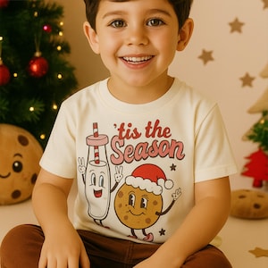 May include: A child's cream-coloured t-shirt with the text "'tis the Season" in red and pink. The shirt features a cartoon milk and cookie design with a Santa hat. The child is smiling in front of a Christmas tree.