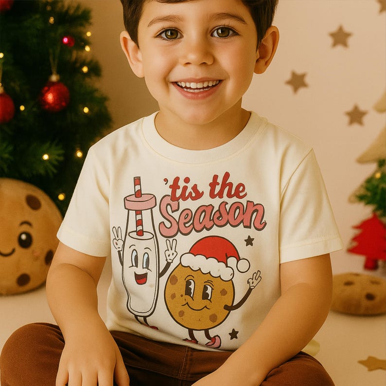 May include: A child's cream-coloured t-shirt with the text "'tis the Season" in red and pink. The shirt features a cartoon milk and cookie design with a Santa hat. The child is smiling in front of a Christmas tree.