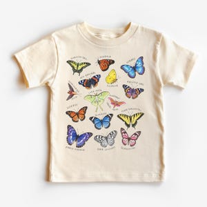 May include: Cream-colored short-sleeved t-shirt featuring a colorful butterfly graphic. The graphic includes various butterfly species with their names, such as Monarch, Swallowtail, and Purple Emperor. The shirt is made of a soft material.