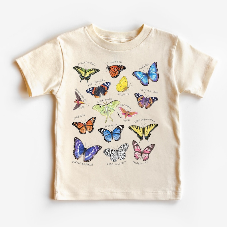 May include: Cream-colored short-sleeved t-shirt featuring a colorful butterfly graphic. The graphic includes various butterfly species with their names, such as Monarch, Swallowtail, and Purple Emperor. The shirt is made of a soft material.