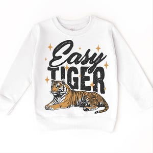 Op de afbeelding: Witte longsleeve trui met de tekst "Easy Tiger" in zwart met gouden sterren. Een cartoon tijger ligt onder de tekst.