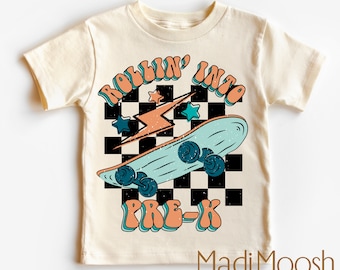 Puede incluir: Una camiseta de color crema con un gráfico de una patineta, un rayo y estrellas sobre un fondo a cuadros blanco y negro. El texto "Rollin' into Pre-K" está en una fuente retro. El texto "Madi Moosh premium clothing MADE WITH LOVE" está en la parte inferior de la camiseta.