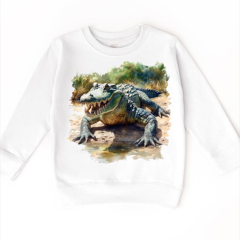 K&ouml;nnte beinhalten: Wei&szlig;er Pullover mit einem gr&uuml;nen Alligator-Grafikdruck. Der Alligator befindet sich in einer sumpfigen Umgebung mit gr&uuml;nem Laub und Wasser.