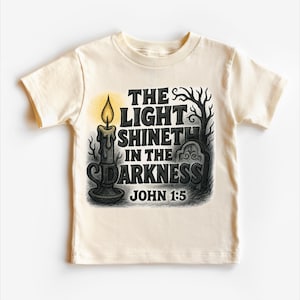 May include: Cream-colored t-shirt featuring a graphic with a lit candle, a tombstone, and the text "THE LIGHT SHINETH IN THE DARKNESS JOHN 1:5" in black. The design includes a tree silhouette and a glowing yellow flame.
