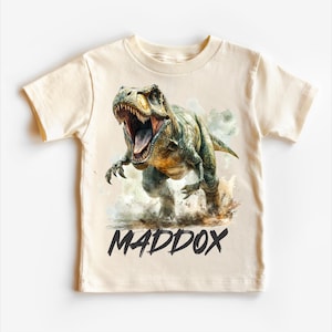 May include: A beige t-shirt with a graphic of a green dinosaur roaring. The dinosaur is in a watercolor style with a brown background. The text "MADDOX" is printed in black below the dinosaur.