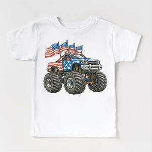Pode incluir: Camiseta branca com uma ilustra&ccedil;&atilde;o de desenho animado de um monster truck. O caminh&atilde;o &eacute; decorado com a bandeira americana e tem pneus pretos grandes. V&aacute;rias bandeiras tremulando criam um tema patri&oacute;tico.