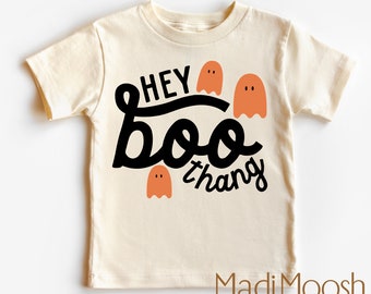 Puede incluir: Camiseta color crema con la frase "Hey Boo Thang" en escritura negra y tres fantasmas naranjas. Es una camiseta de manga corta y cuello redondo, perfecta para Halloween. La marca "Madi Moosh" está impresa en la parte inferior.