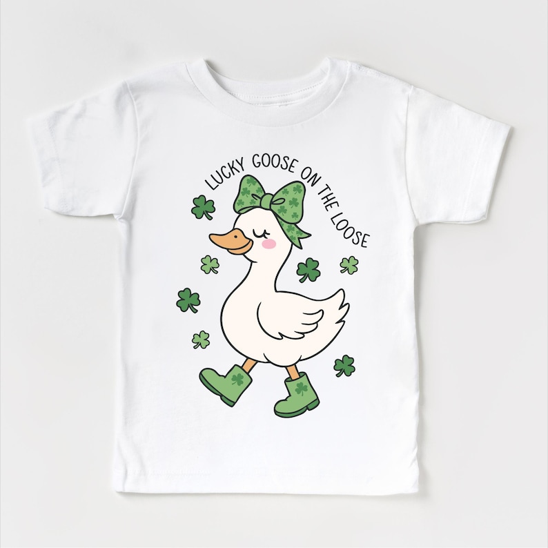 May include: White t-shirt featuring a cartoon goose with a green bow, boots, and shamrocks. The text "LUCKY GOOSE ON THE LOOSE" is arched above the goose. The design is perfect for St. Patrick's Day.