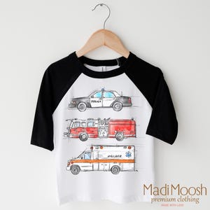 May include: White t-shirt with black raglan sleeves featuring a watercolor illustration of a police car, fire truck, and ambulance. The text "POLICE" is visible on the police car. The text "MADI MOOSH premium clothing MADE WITH LOVE" is visible at the bottom of the shirt.