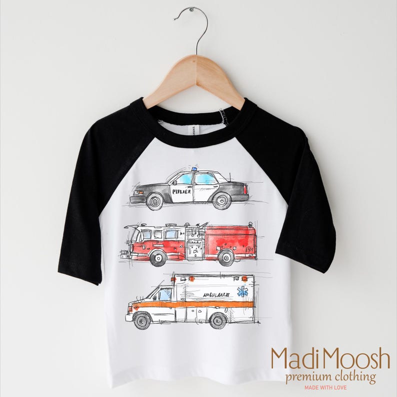 May include: White t-shirt with black raglan sleeves featuring a watercolor illustration of a police car, fire truck, and ambulance. The text "POLICE" is visible on the police car. The text "MADI MOOSH premium clothing MADE WITH LOVE" is visible at the bottom of the shirt.