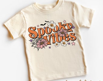 Puede incluir: Camiseta color crema con las palabras "Spooky Vibes" en naranja, rodeada de flores, estrellas, una telaraña y murciélagos. La camiseta es de manga corta y cuello redondo.