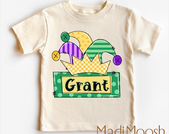 Può includere: Una maglietta color crema con un designo di cappello da giullare verde, giallo, viola e nero. Il cappello è posto su una scatola verde con pois bianchi e il nome "Grant" scritto in nero.