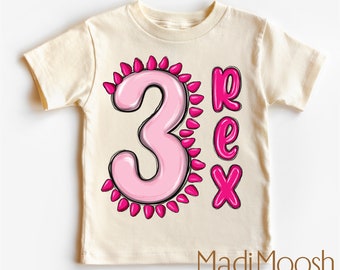 May include: A light beige toddler t-shirt with a pink number 3 surrounded by pink spikes and the word "Rex" in pink letters. The shirt has the text "Madi Moosh premium clothing MADE WITH LOVE" at the bottom.