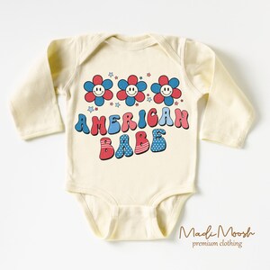 May include: Cream-colored baby bodysuit with long sleeves. The front features the text "AMERICAN BABE" in red, white, and blue, along with smiling flower graphics and stars. The bodysuit is made of soft material.